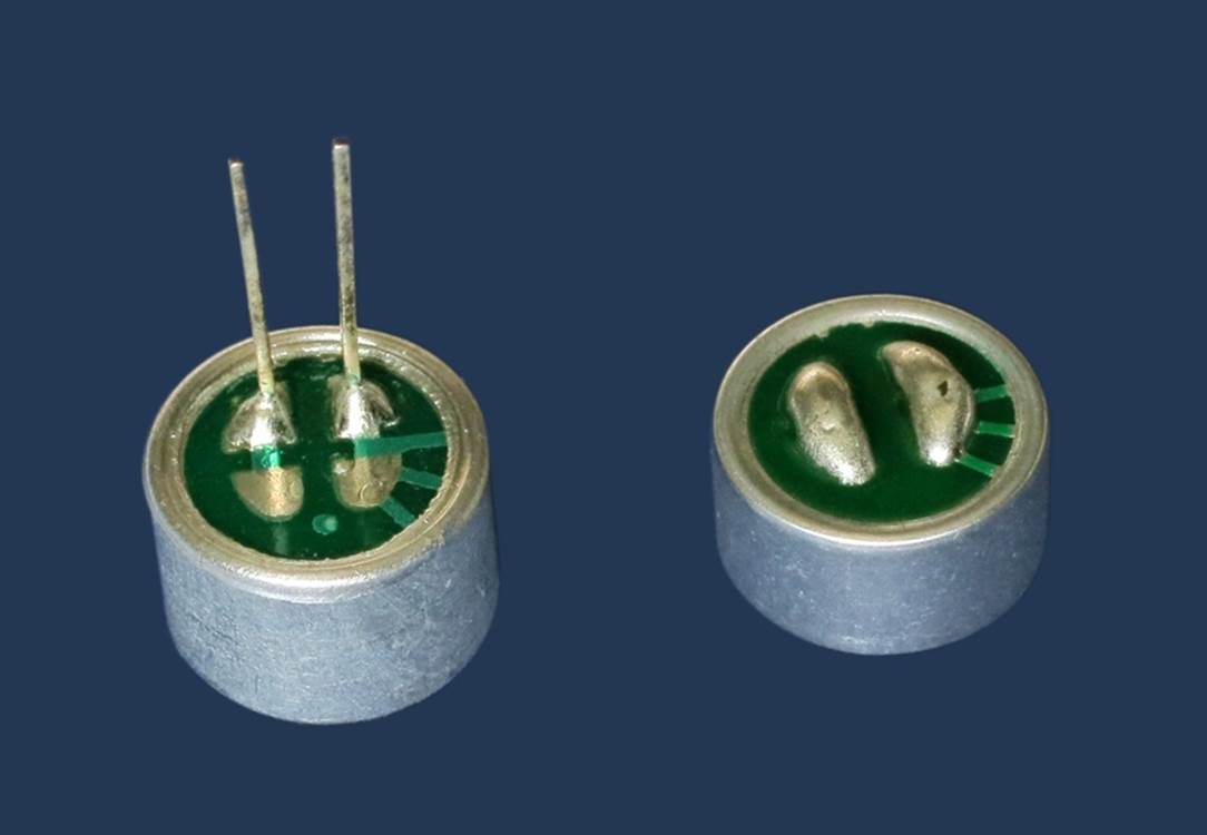 The undersides of two typical electret microphones, one with leads, the other with solder pads for surface-mount. The ground terminal is on the right in each case, as indicated by the metal “fingers” visible through the green translucent insulating layer.